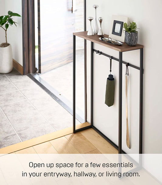 A black metal and MDF console table with open shelves, displaying various items such as a plant, a book, and a yoga mat, set in an entryway or hallway of a home.