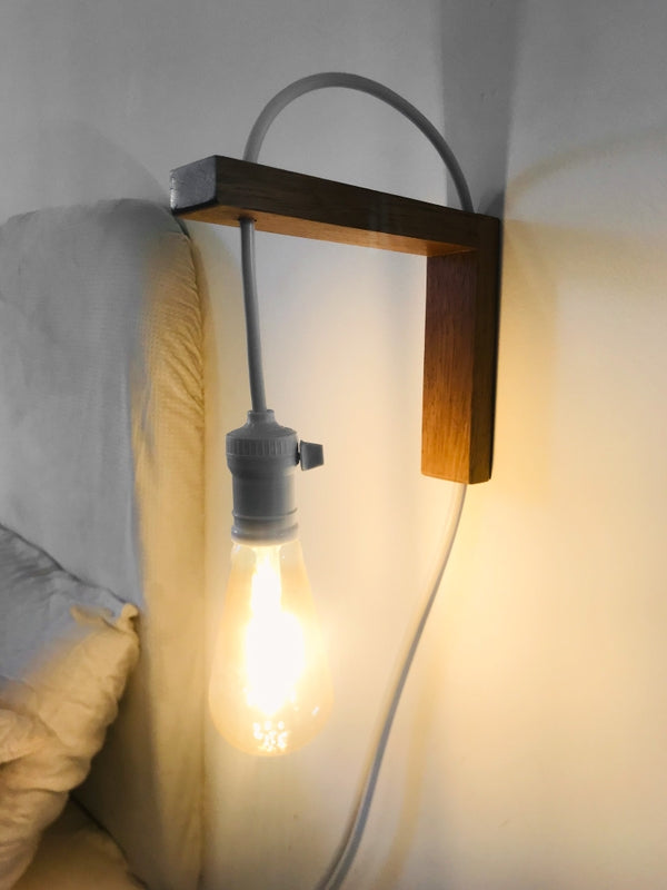 A wall-mounted bedside lamp with a wooden structure and a glowing bulb, installed next to a pillow on a bed.
