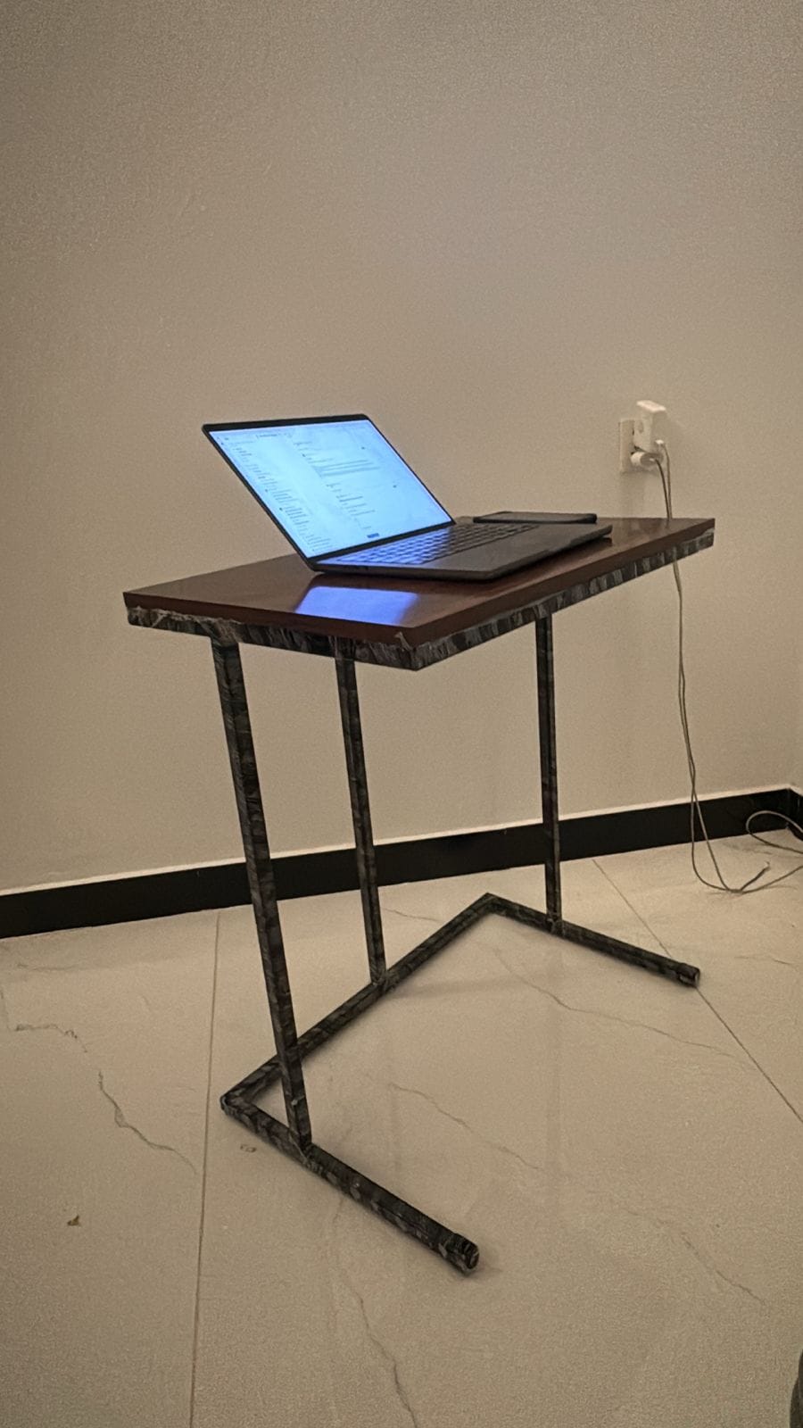 A dark wood and black metal standing desk with an open laptop on top, set in a room with a beige wall and tiled floor.